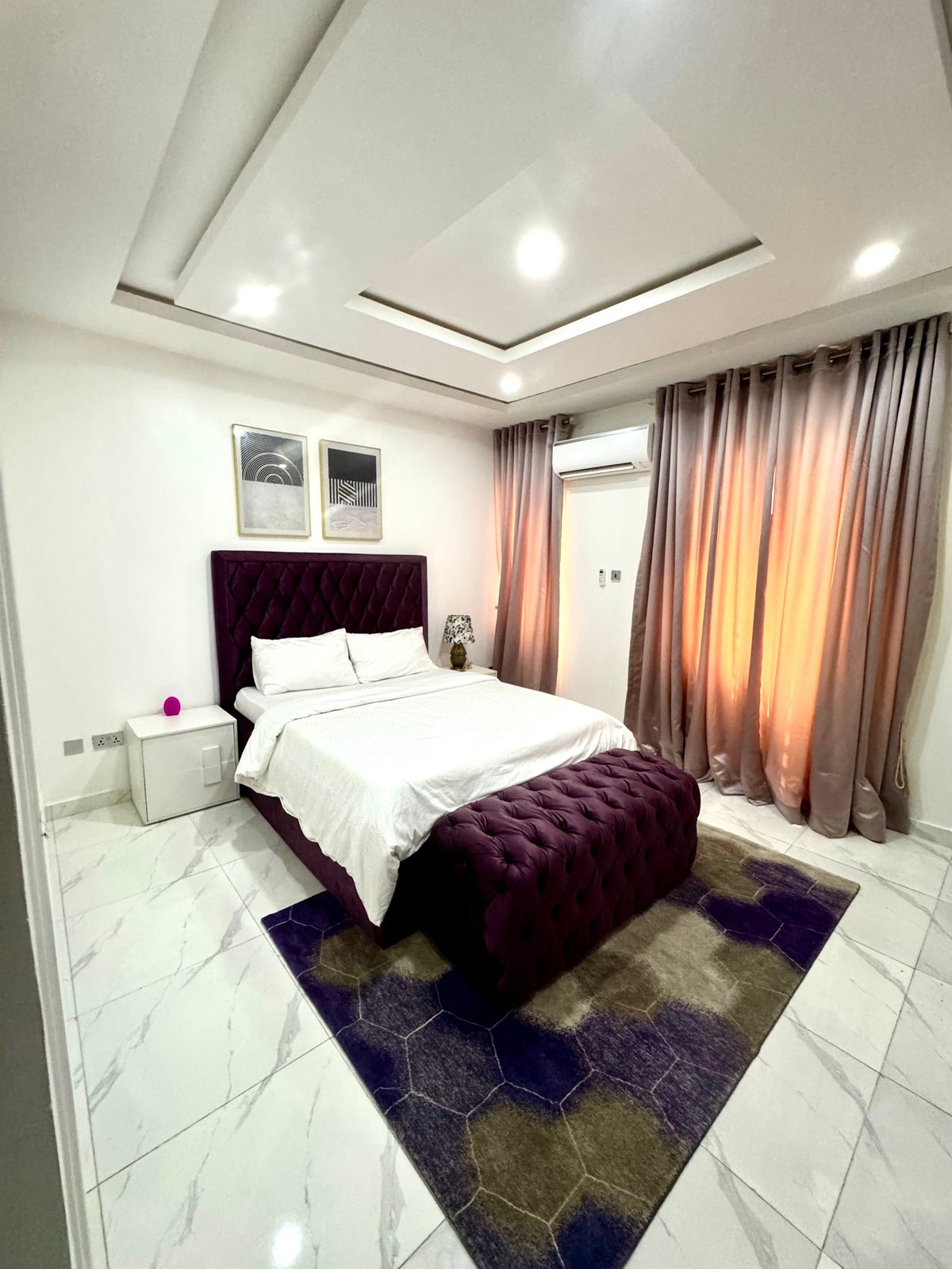 Bedroom with purple velvet bed, white bedding, geometric area rug and floor-to-ceiling curtains